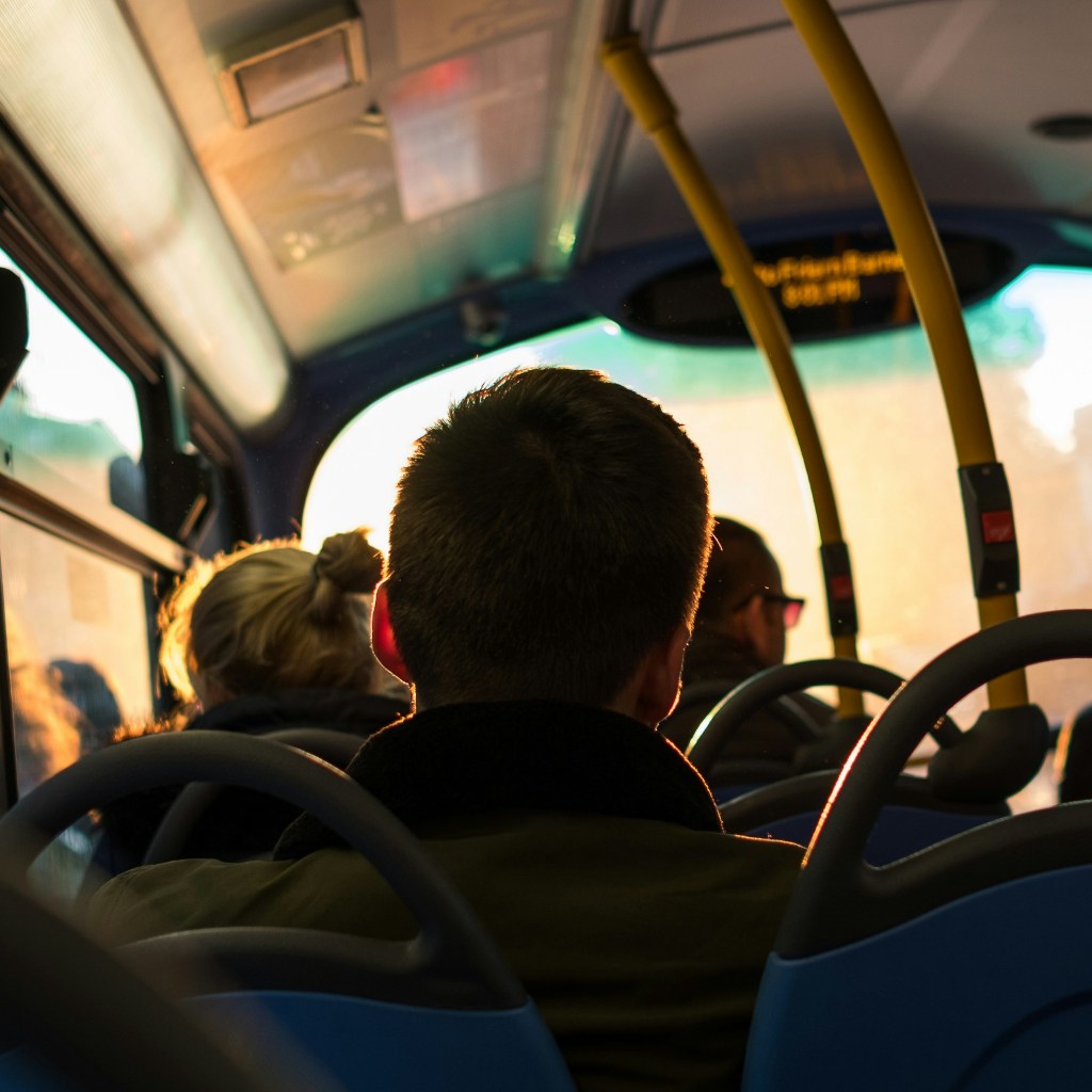 Interior of a public bus during sunset, with passengers seated facing forward and warm sunlight streaming through the windows. Yellow handrails and blue seatbacks are visible, and a digital display is mounted at the front.