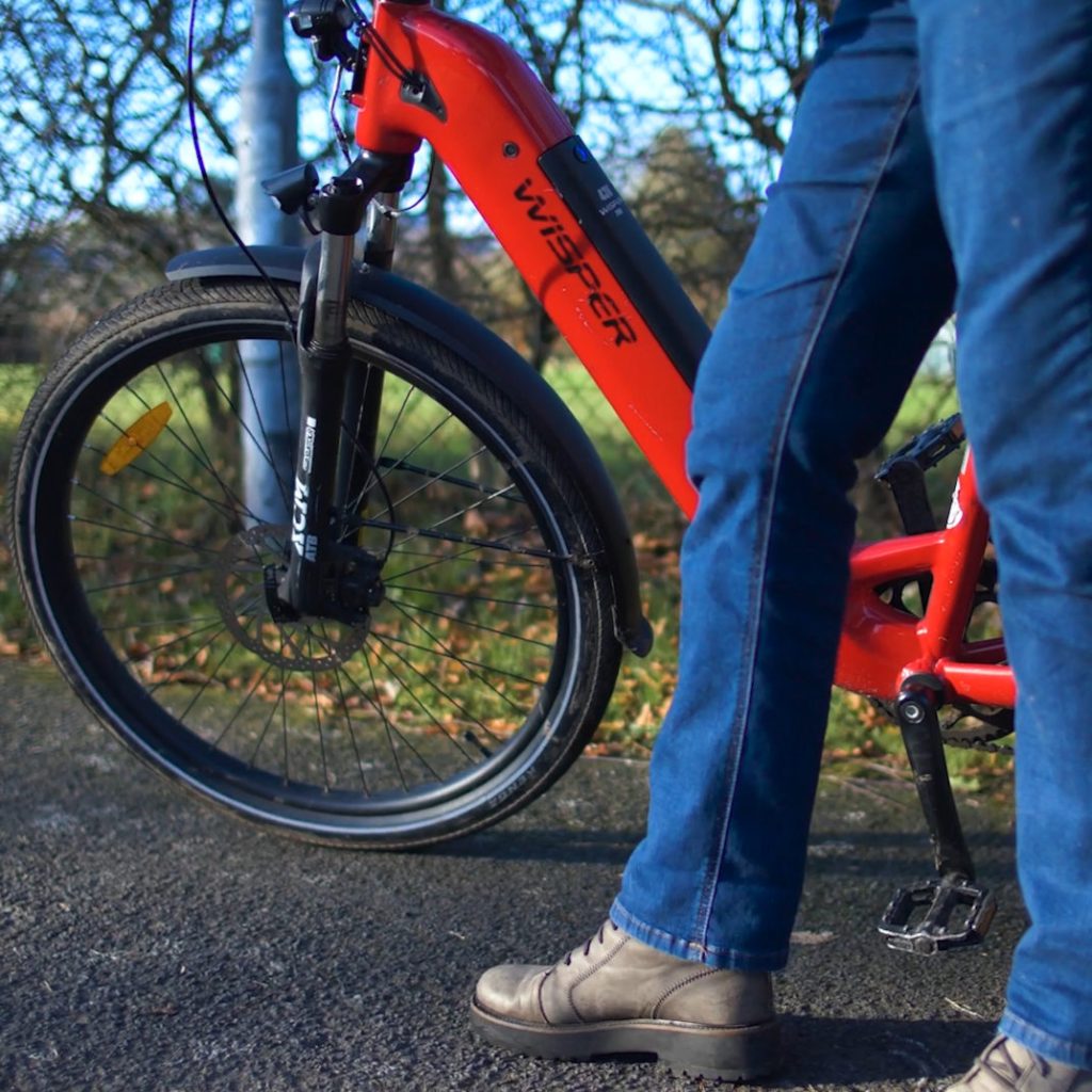 A close-up view shows a person standing beside a red Wisper‑branded electric bicycle on a paved path. The bike’s front wheel and suspension fork, labelled ‘RST AERIAL’, are prominently visible, emphasising its sturdy design. The person, dressed in blue jeans and grey lace‑up shoes, stands next to the bike as if preparing to ride. Behind them, leafless trees and a patch of winter grass suggest a cold‑season outdoor setting. The image highlights the design and branding of the electric bicycle and conveys its use for recreation or eco‑friendly transport.