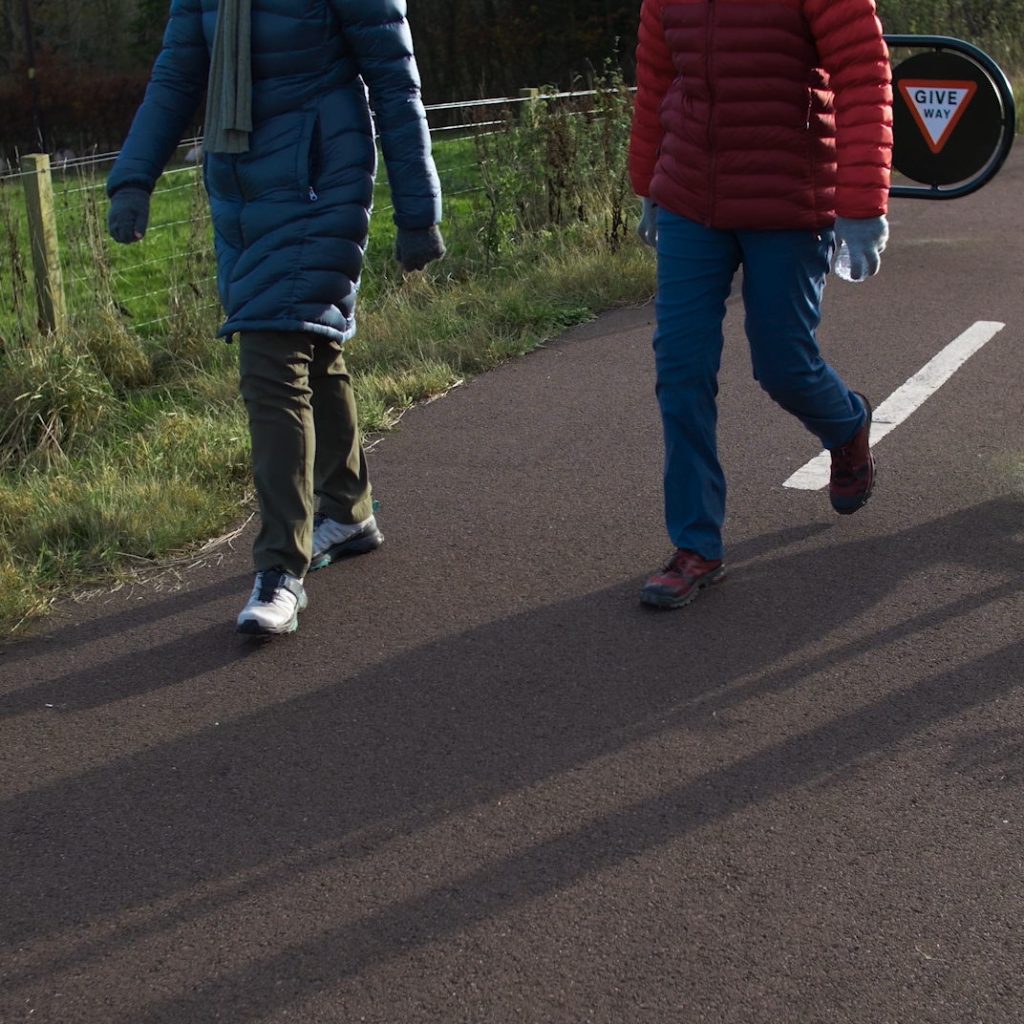 Two people in winter clothing walking along a paved path bordered by grass and a wire fence, with a ‘GIVE WAY’ sign visible in the background. The low sun casts long shadows, suggesting a cold, clear day.