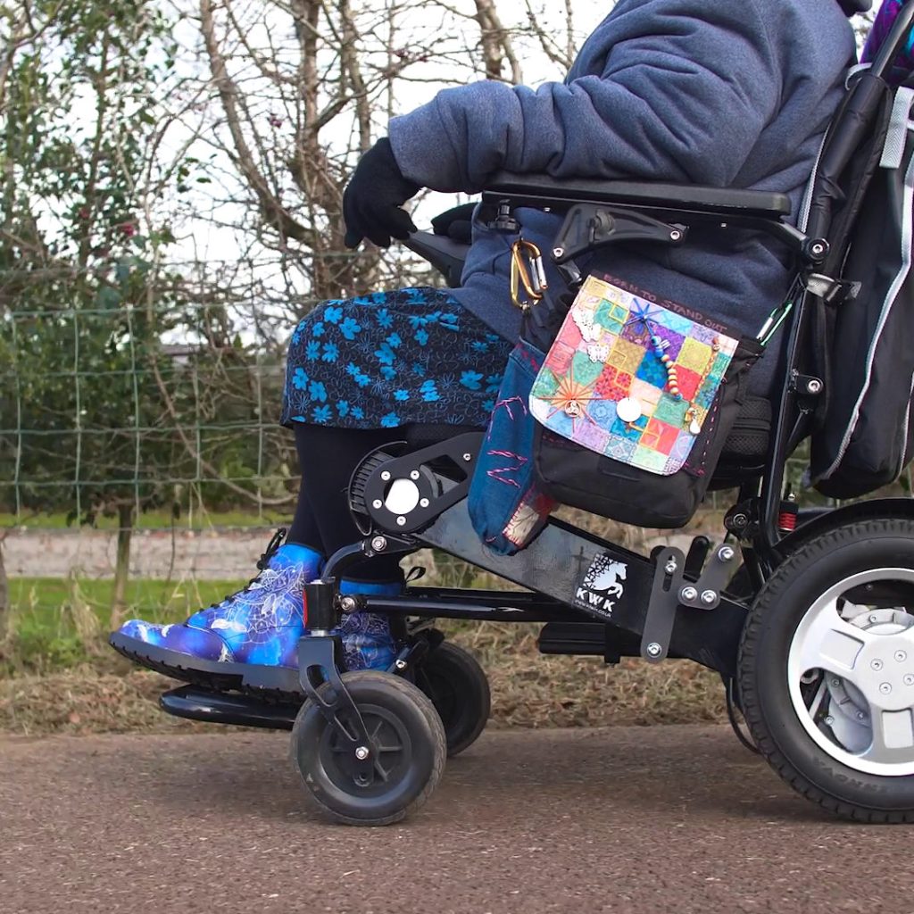 A person sits outdoors in a motorised wheelchair on a paved path, dressed warmly in a blue jacket, patterned blue‑and‑black trousers, and blue boots with a white web‑like design. The wheelchair features large rear wheels, smaller front wheels, visible mechanical components, and branding that includes a horse logo and the letters ‘KWK’. A colourful patchwork bag hangs from the side of the chair, adding a personalised touch. Behind the person, a wire fence and leafless trees indicate a winter or early‑spring setting. The image highlights mobility support, individuality, and outdoor accessibility.