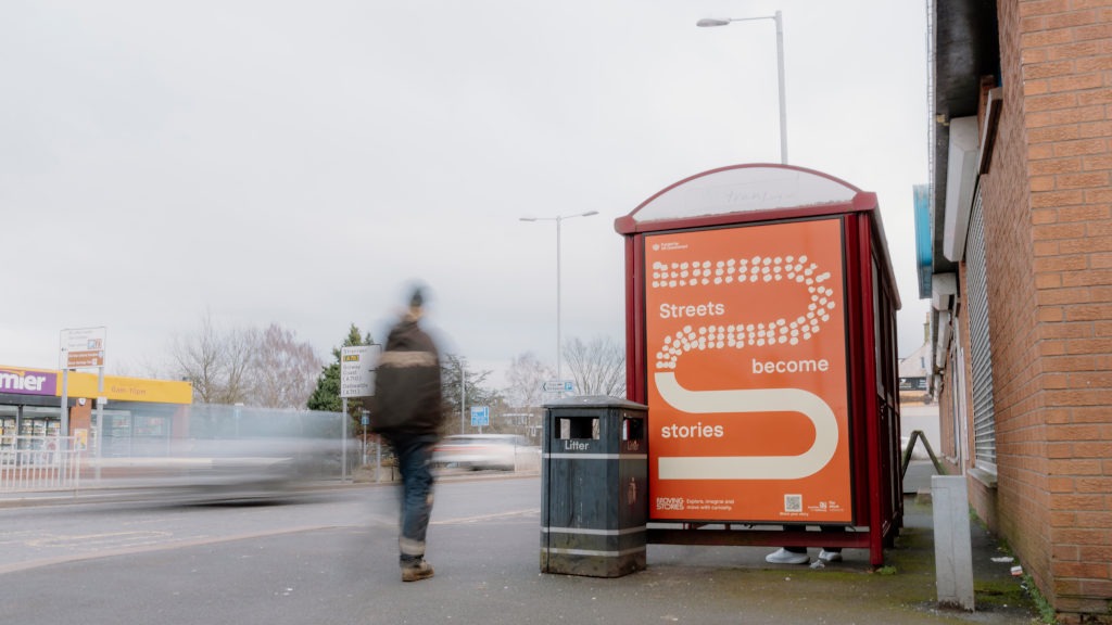 A bus stop on Glasgow Street in Dumfries features a bright orange poster with a stylised white dotted path forming an S‑shape and the words ‘Streets Become Stories’ integrated into the design. The poster includes a QR code and an invitation to explore, imagine and share personal stories. A litter bin marked ‘Litter’ stands in the foreground, while a passer‑by walks past in motion blur. Behind the shelter, a Premier convenience store and blurred traffic contribute to a lively town scene, emphasising everyday movement and the campaign’s focus on local storytelling.