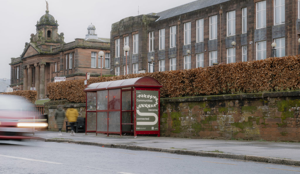 A historic stone building, Dumfries Academy, with classical columns and a domed roof topped by a statue stands behind a busy street scene. To the right, a bright red bus‑stop shelter displays a poster reading ‘Communities Become Connected’. A person in a yellow coat walks past the shelter, while a vehicle on the left appears blurred in motion, suggesting passing traffic. Trimmed hedges and low stone walls frame the area, creating a well‑kept town setting that contrasts the building’s heritage architecture with modern public‑transport infrastructure.